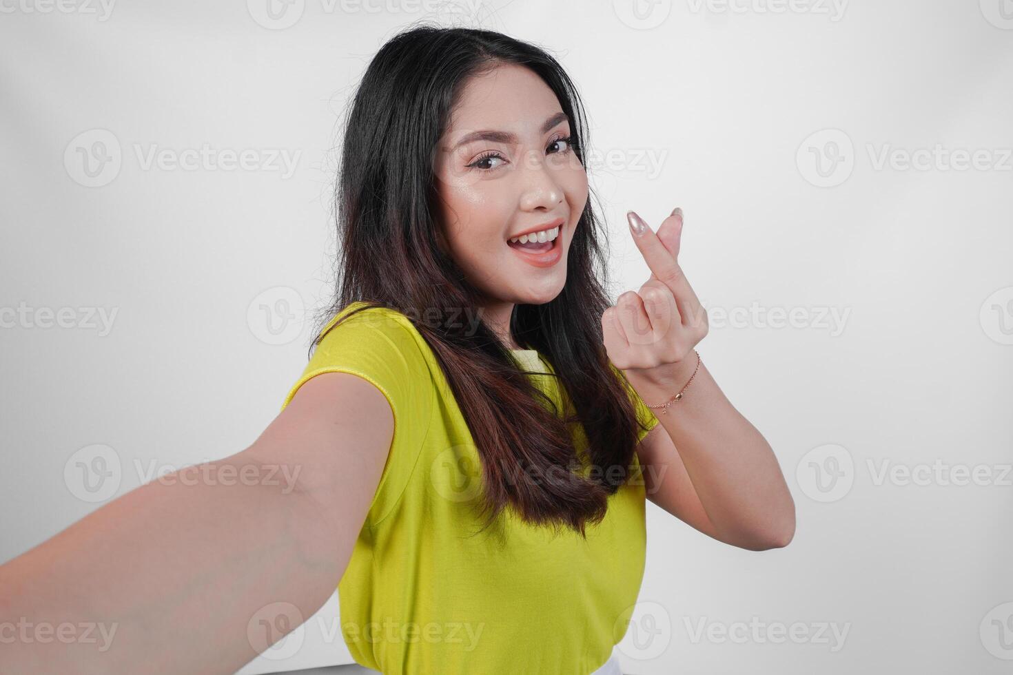 Lovely young Asian woman in a yellow shirt is smiling and showing finger heart gesture to the camera, isolated by white background. photo