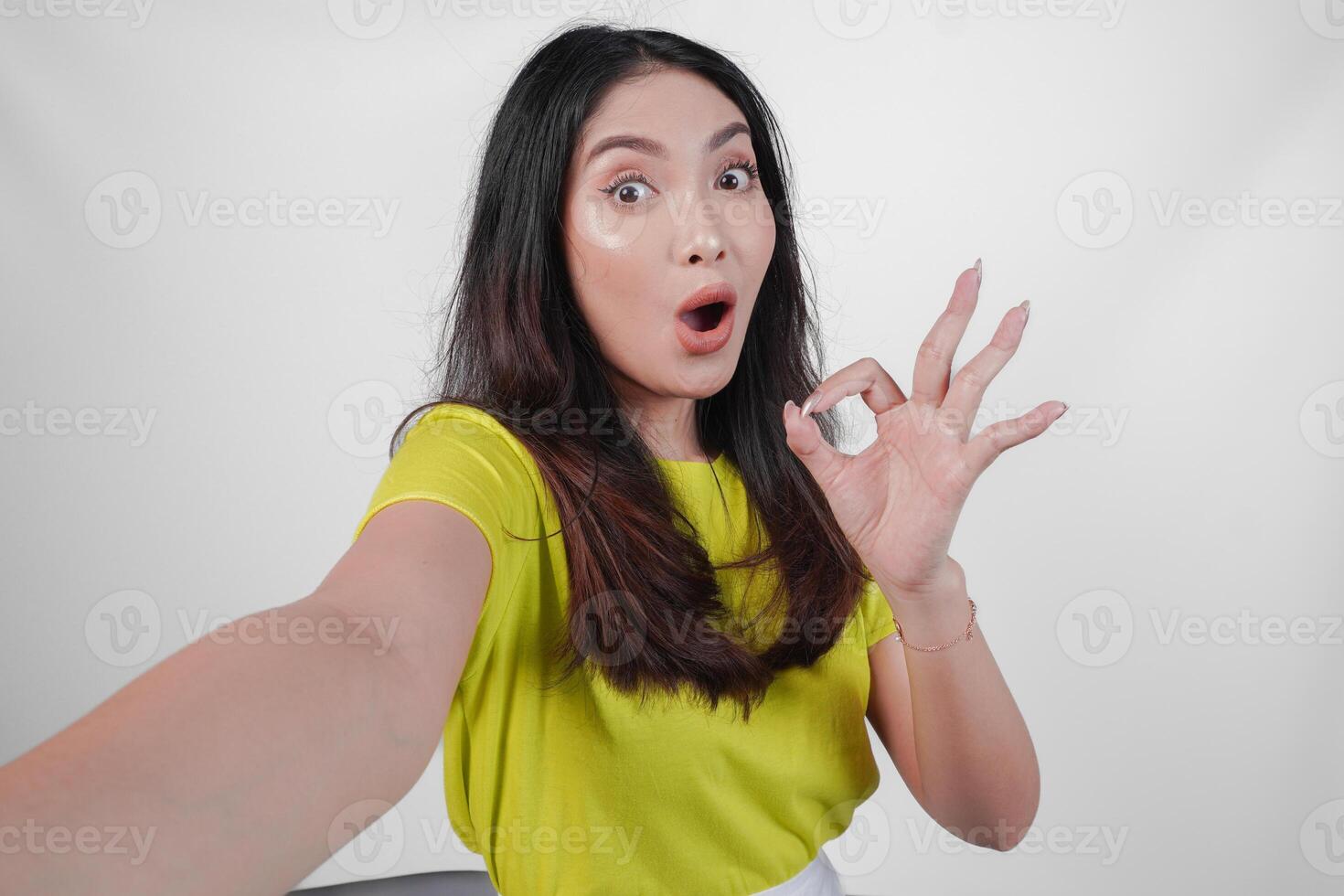 Attractive young Asian woman in a yellow shirt is taking a selfie and giving okay sign. Concept of happiness and positivity, as the woman is smiling and expressing approval. photo