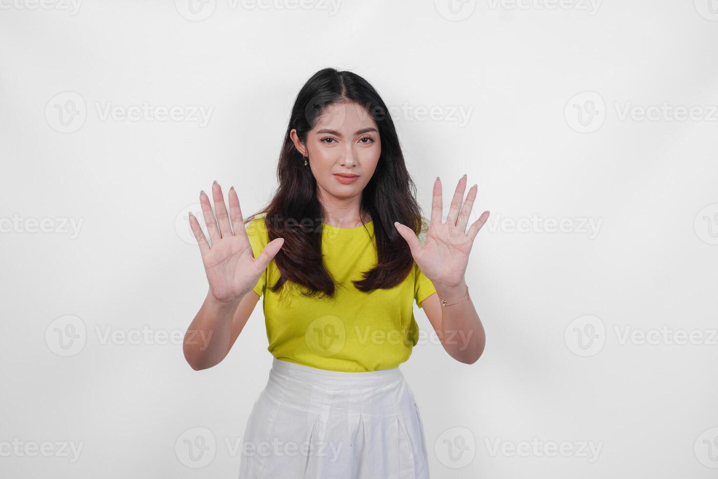 Serious young Asian woman in a yellow shirt and white skirt is holding up her hand to make a stop sign. photo