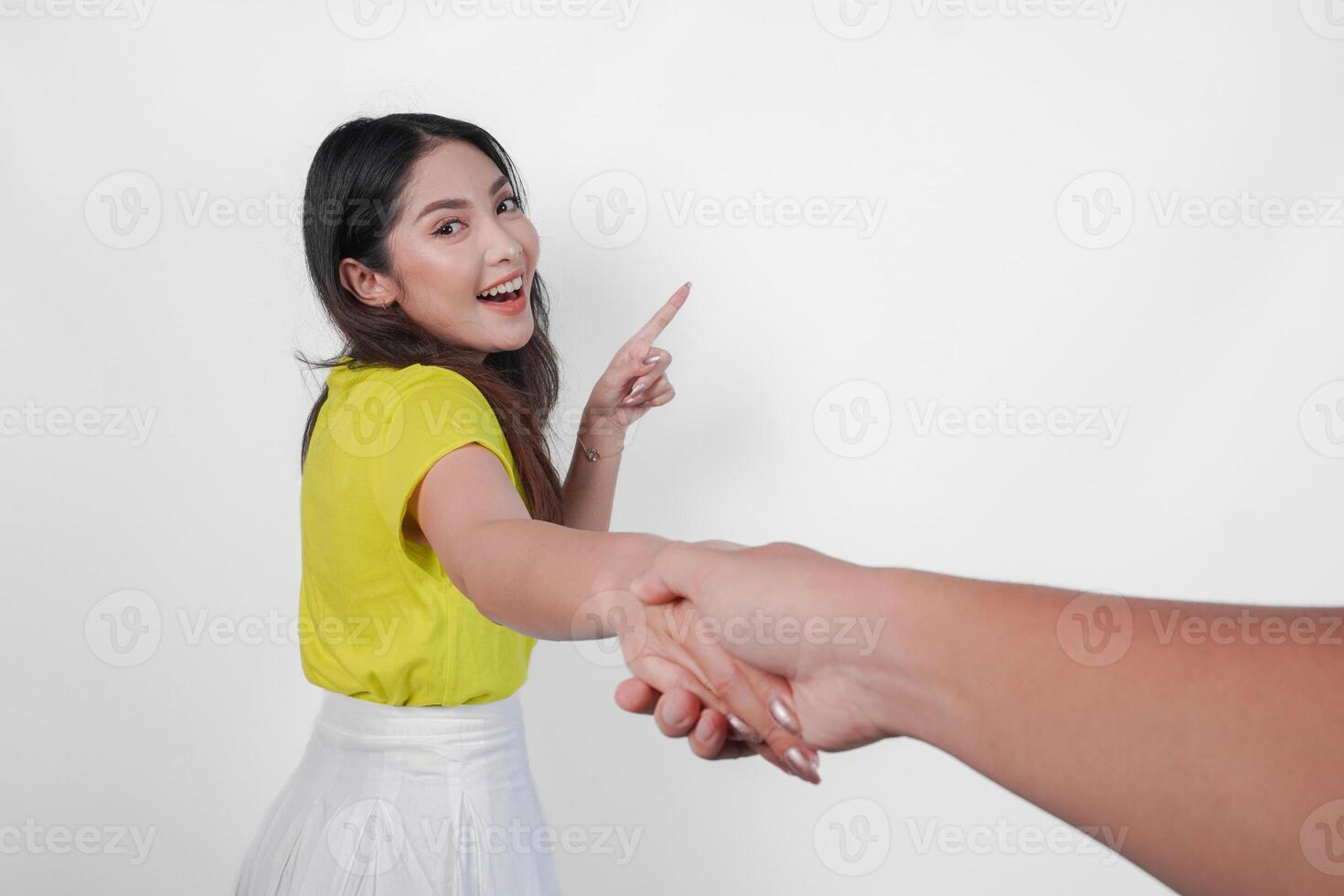 A happy Asian woman in a yellow shirt is holding hands with another person, asking to follow over while pointing to the copy space isolated white background. photo