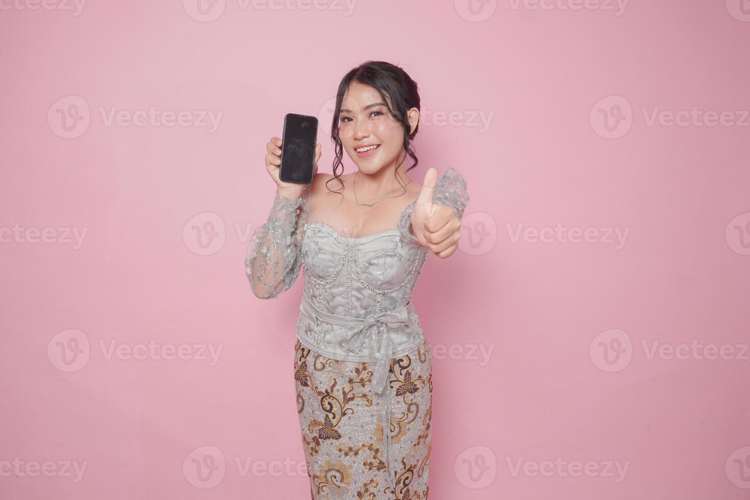 Excited Indonesian woman in modern kebaya dress gives thumbs up hand gesture of approval while showing copy space on her phone, isolated by pink background. photo