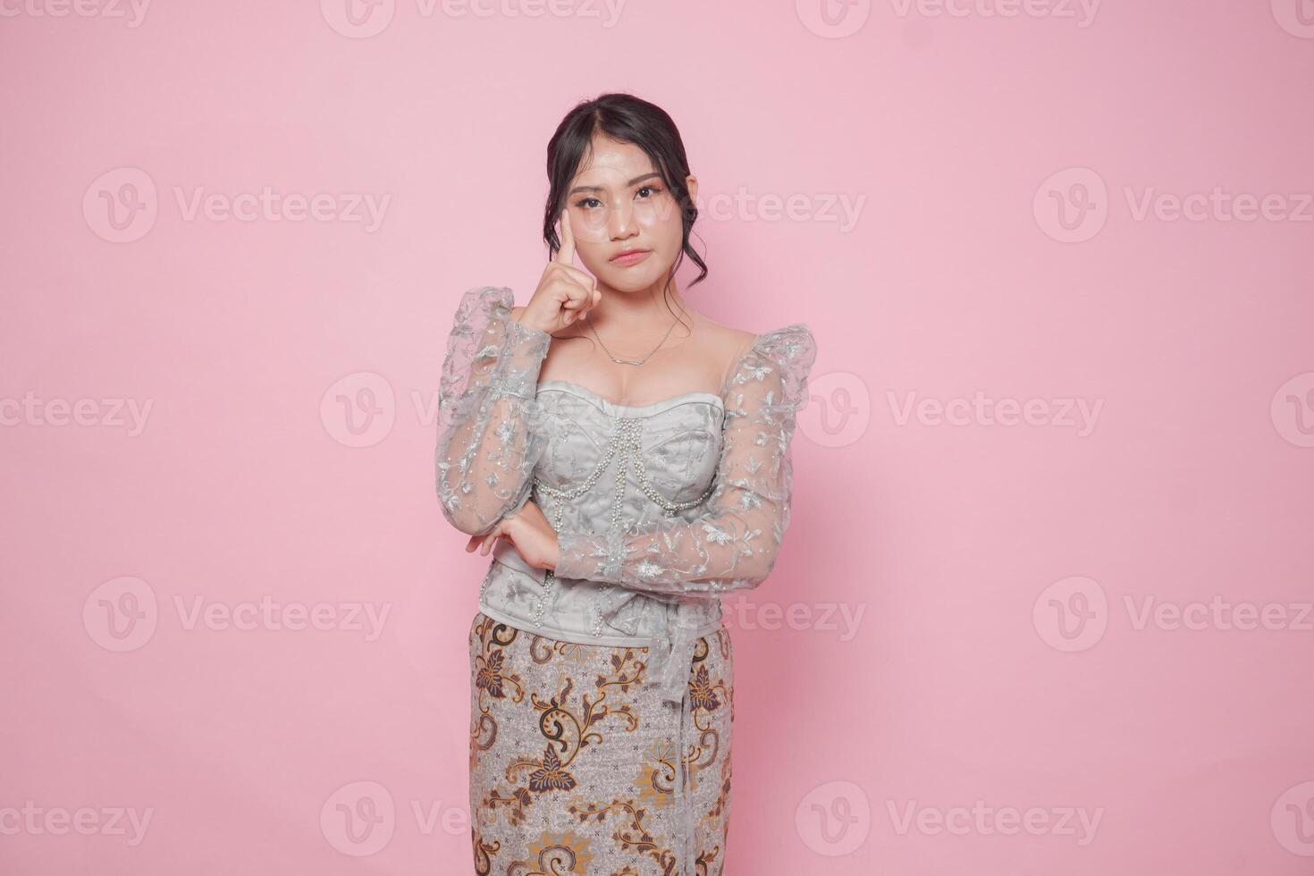 Thoughtful Asian woman wearing modern kebaya dress tilting her head, isolated by pink background. photo