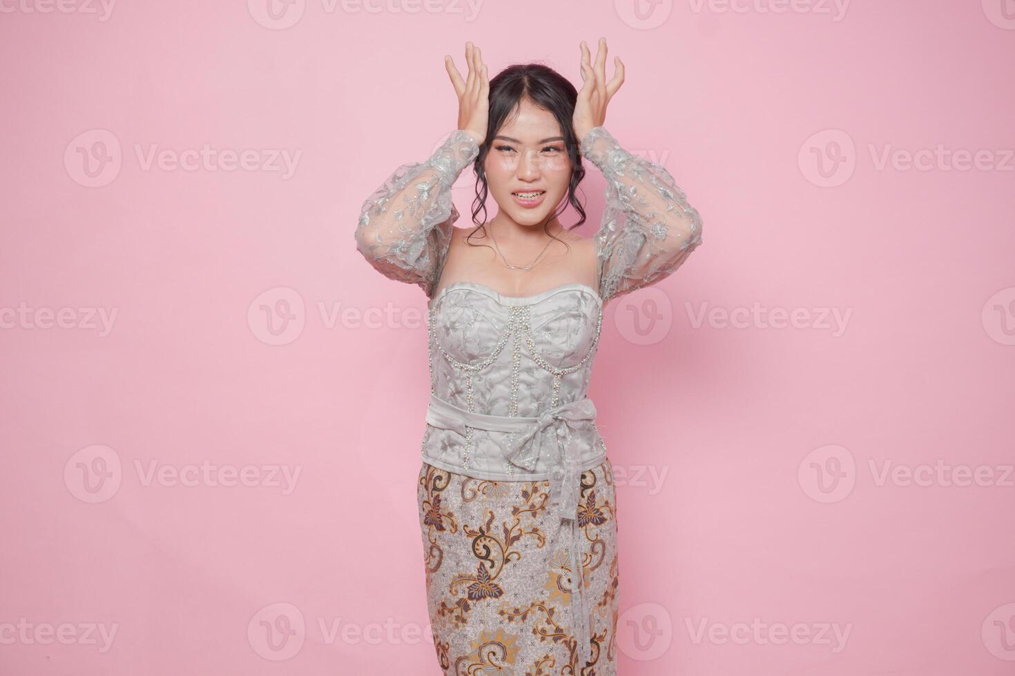 Young Asian woman wearing modern kebaya dress looking confused while holding her head, isolated by pink background. photo