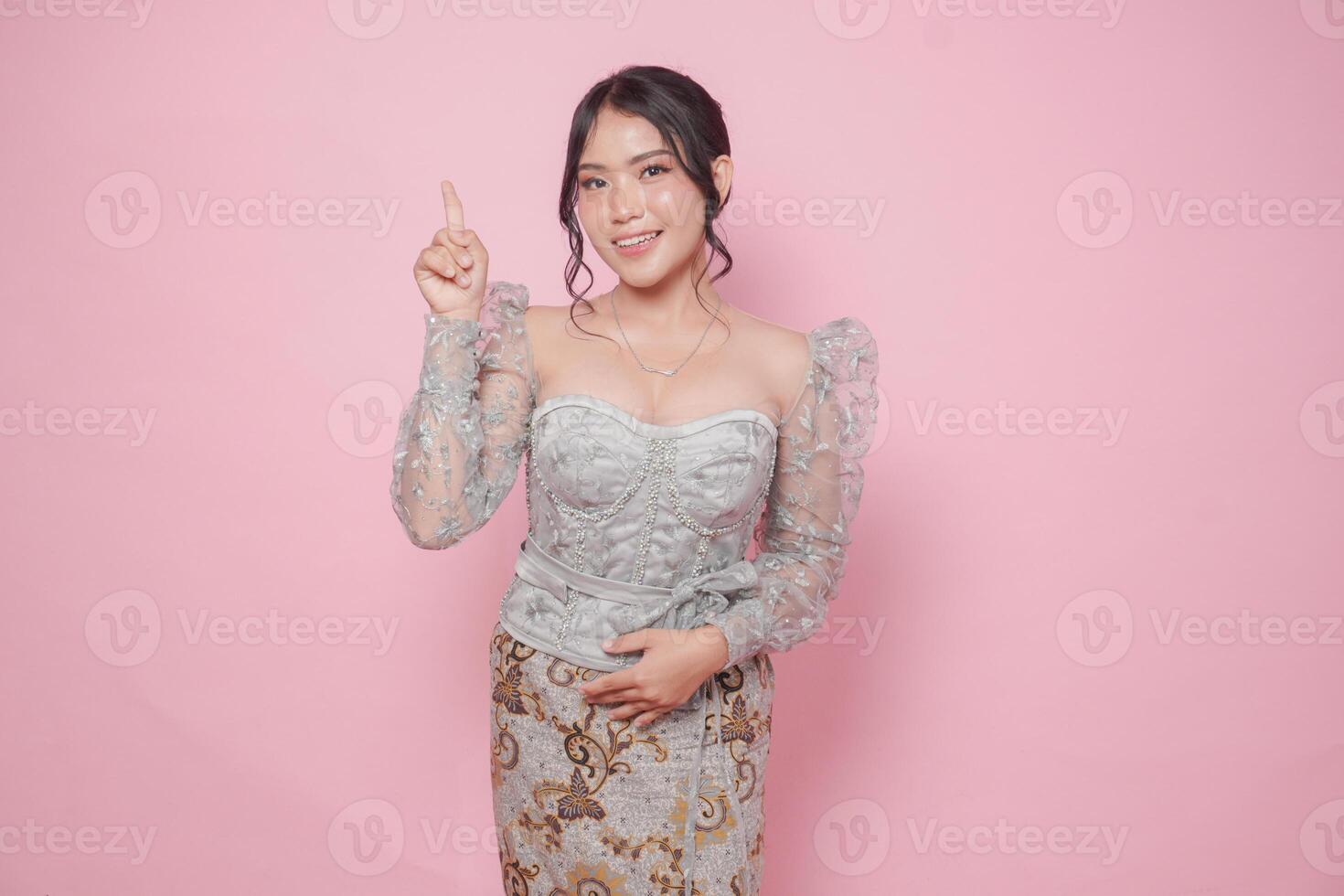 Beatiful Asian woman wearing modern kebaya dress pointing at the copy space upside her, isolated by pink background. photo
