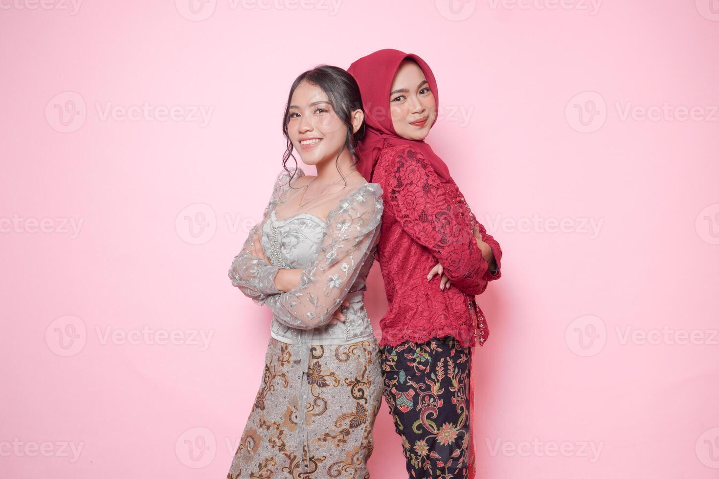 Two smiling Asian women wearing modern kebaya dress posing and leaning on each other back, isolated by pink background. photo