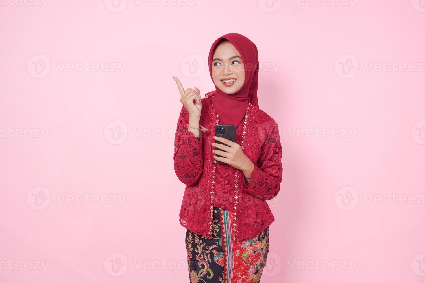 Joyful Asian woman wearing modern kebaya dress and hijab pointing at the copy space upward while holding smartphone, isolated by pink background. photo