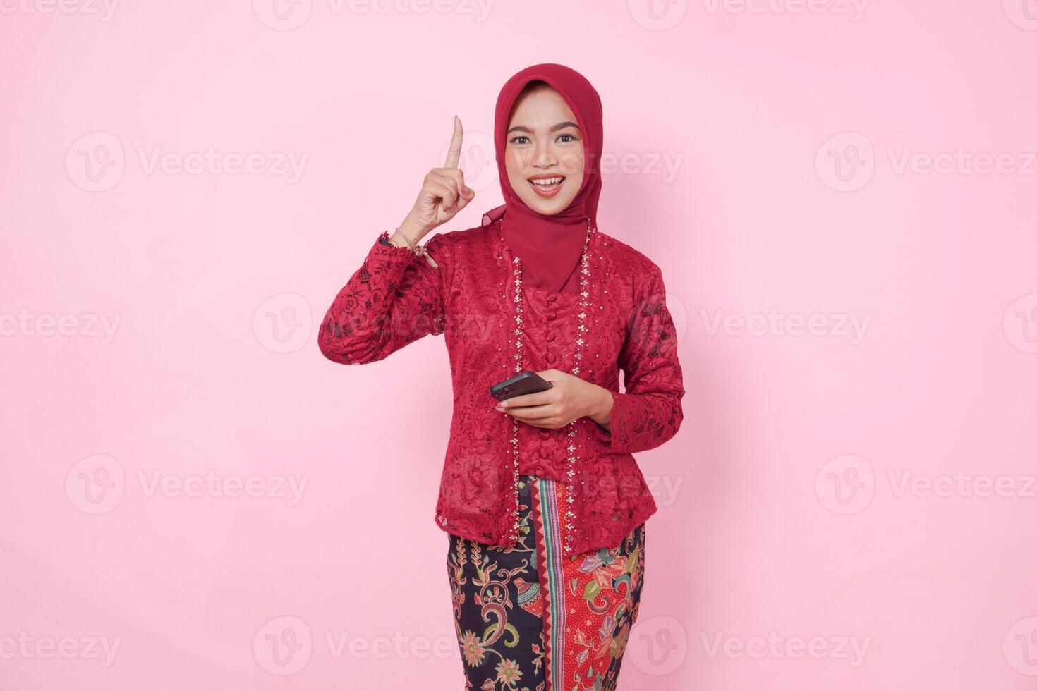 Joyful Asian woman wearing modern kebaya dress and hijab pointing at the copy space upward while holding smartphone, isolated by pink background. photo
