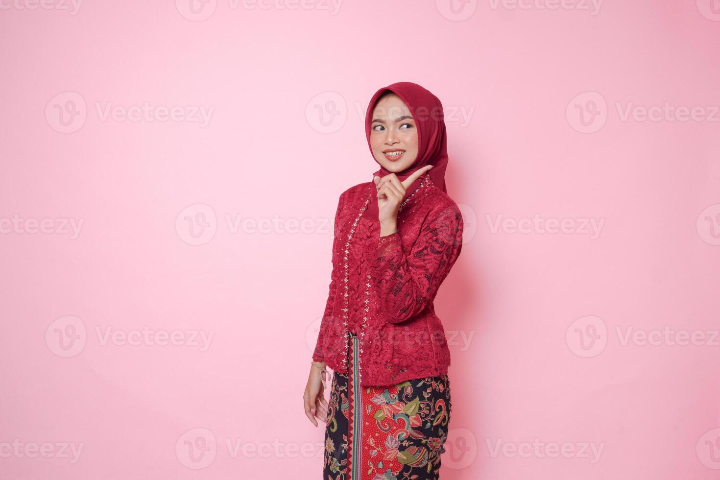 Elegant Asian woman wearing modern kebaya dress and hijab pointing to the copy space behind her back, isolated by pink background. photo