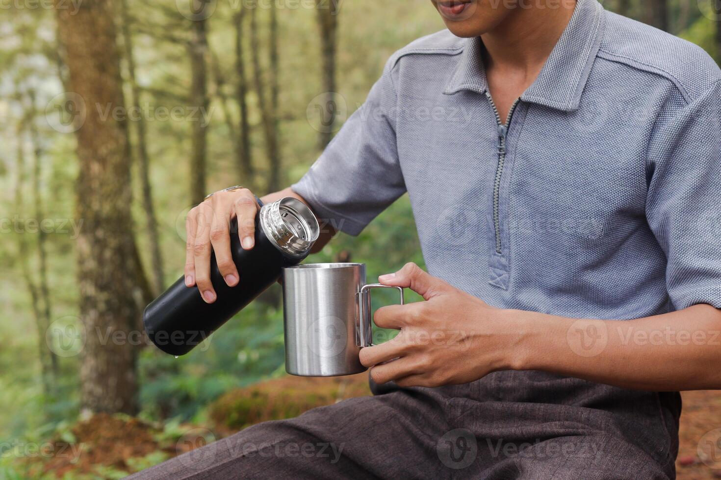 Close up portrait of excited Asian man pouring water from the vacuum flask to the stainless steel mug. Drinking mineral water while trekking in nature forest photo
