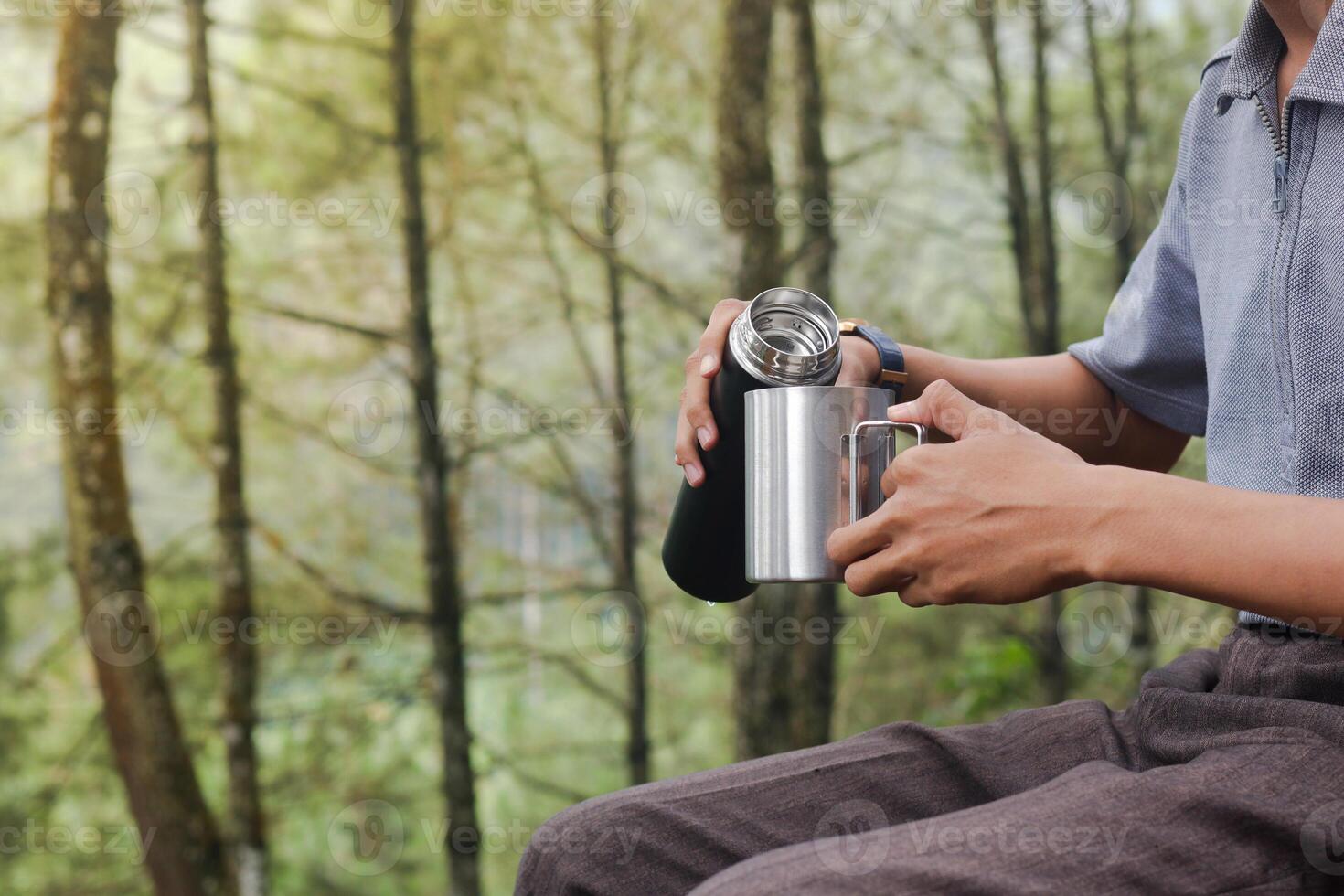 Close up portrait of excited Asian man pouring water from the vacuum flask to the stainless steel mug. Drinking mineral water while trekking in nature forest photo