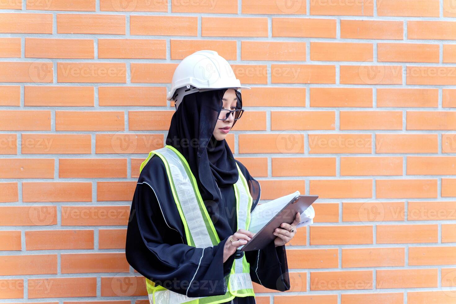 A beautiful Asian Muslim construction engineer portrait in safety vest and Abaya niqab traditional clothes standing and use tablet computer on brick block building wall background. photo