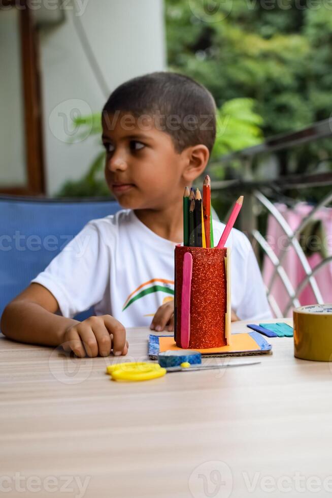 Smart Indian little boy performing best out of waste activity with different colourful objects during the summer vacations, Cute Indian Kid doing best our of waste activity at home on wooden table photo