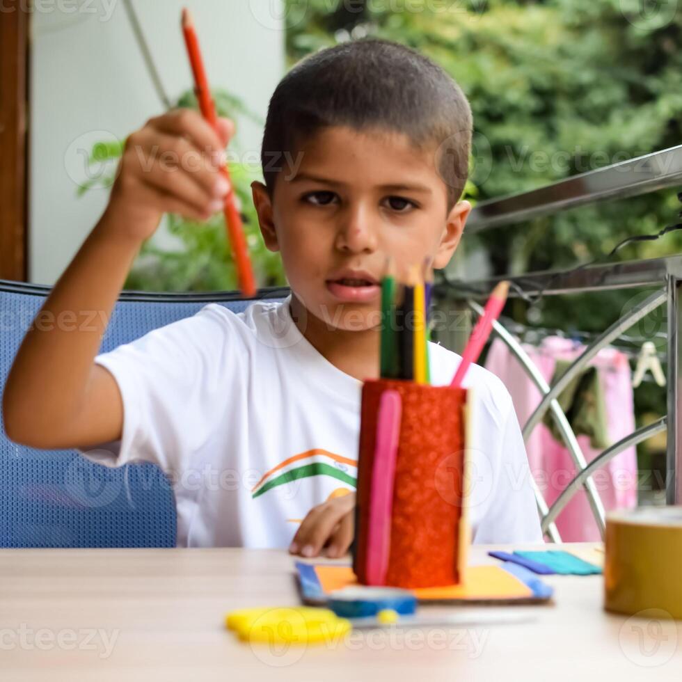 Smart Indian little boy performing best out of waste activity with different colourful objects during the summer vacations, Cute Indian Kid doing best our of waste activity at home on wooden table photo