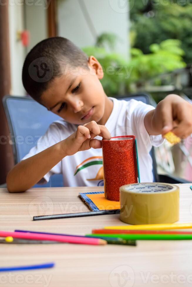 Smart Indian little boy performing best out of waste activity with different colourful objects during the summer vacations, Cute Indian Kid doing best our of waste activity at home on wooden table photo