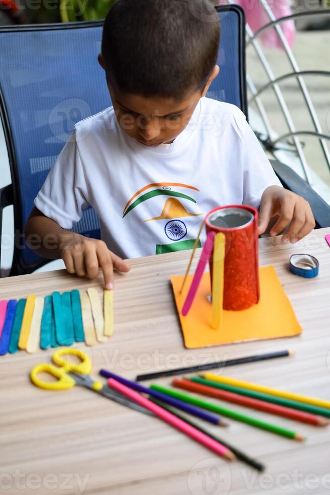 Smart Indian little boy performing best out of waste activity with different colourful objects during the summer vacations, Cute Indian Kid doing best our of waste activity at home on wooden table photo
