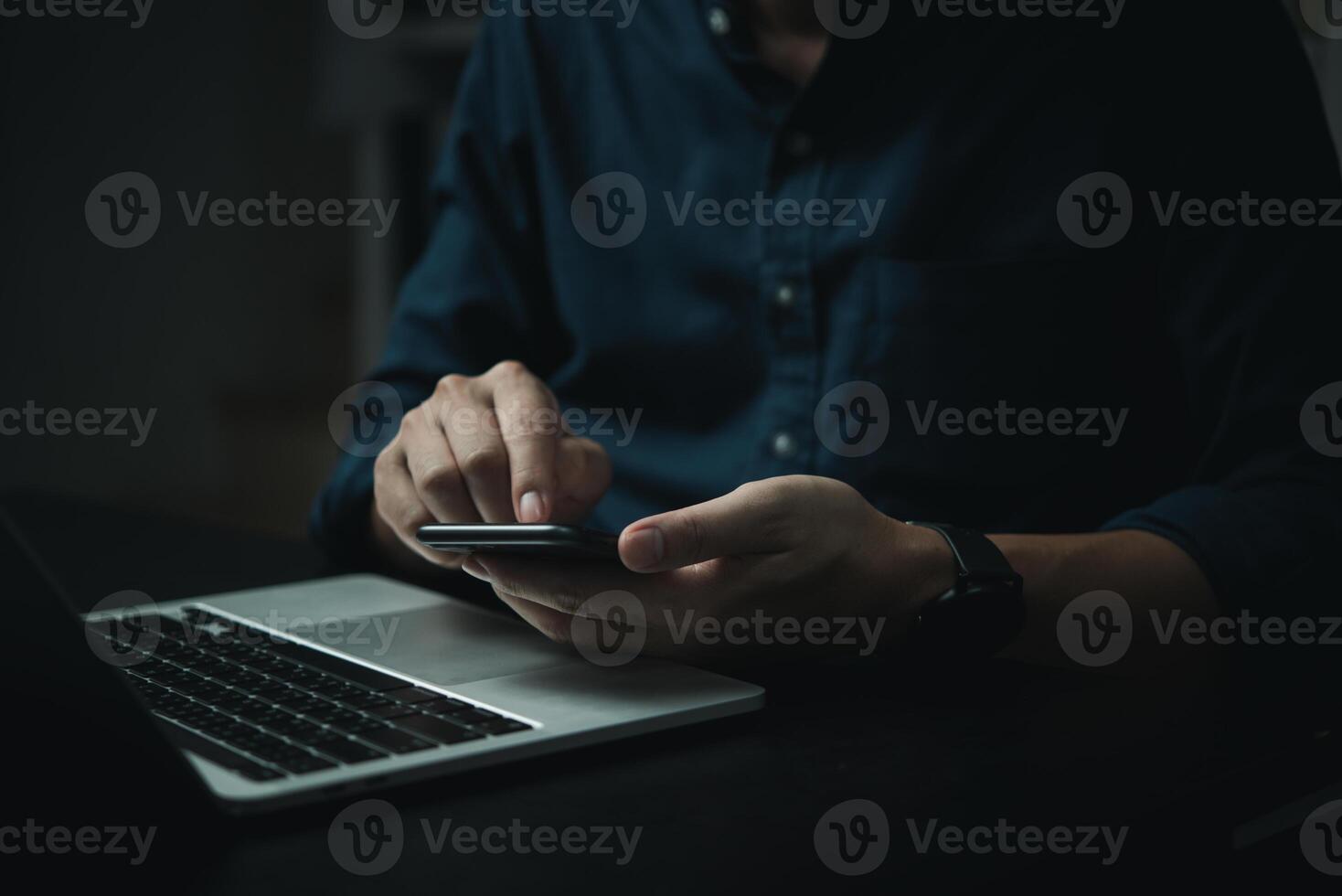 A man is using his cell phone while sitting at a desk with a laptop photo