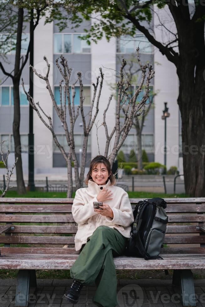 Woman sitting on a park bench smiling while using a smartphone with a backpack. Concept of outdoor relaxation and digital connectivity photo