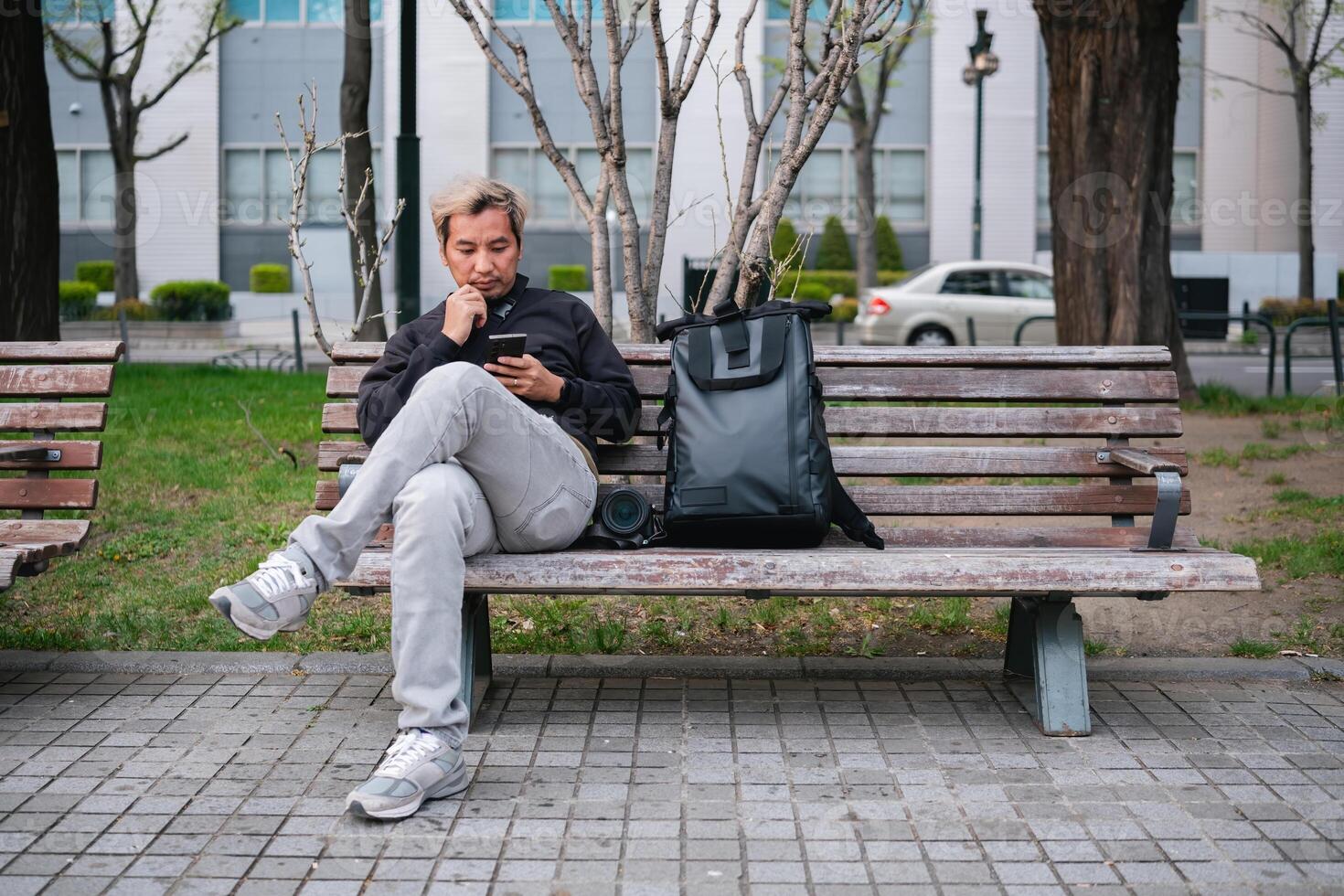 Man sitting on a park bench using smartphone with backpack and camera. Concept of urban relaxation and technology photo