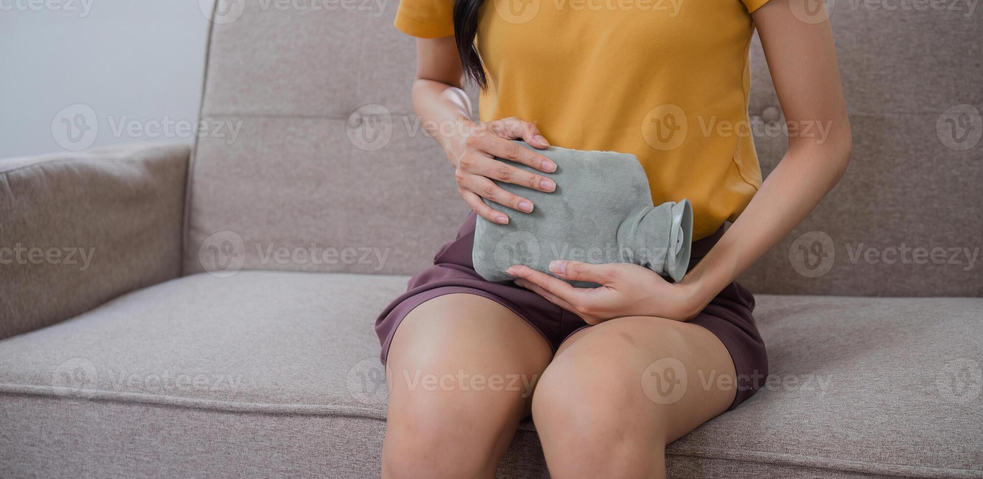 Young Woman Experiencing Menstrual Cramps While Sitting on a Couch, Holding a Hot Water Bottle for Relief photo