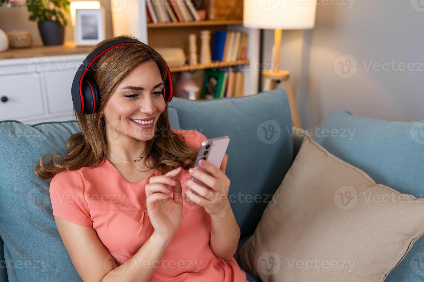 Woman listening music in headphones on windowsill background. Beautiful girl in denim and headphones is listening to music using a smart phone and smiling while lying on bed at home photo