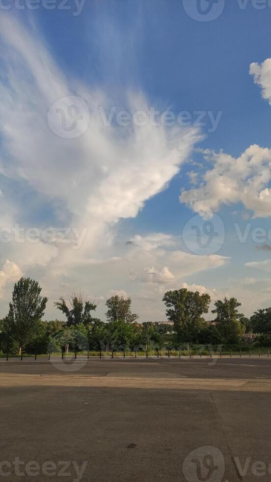 A blue sky with fluffy white clouds above a row of green trees and an empty parking lot photo