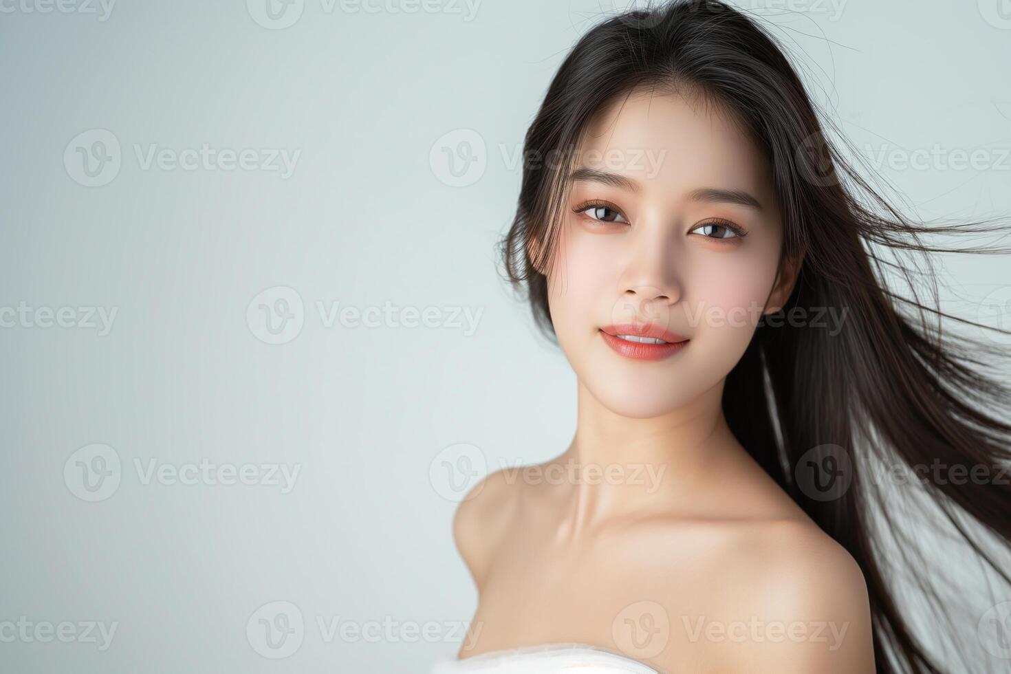 Beautiful Asian woman smiling and touching her face with hands, sitting at a white table on a light grey background, skin care concept photo