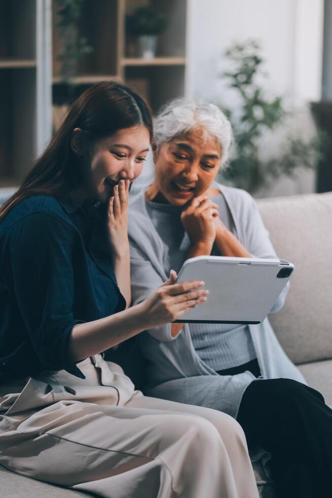Happy grownup daughter showing content on tablet to mature mother. Two family generations women with digital computer gadget resting on couch together, watching videos, making call photo
