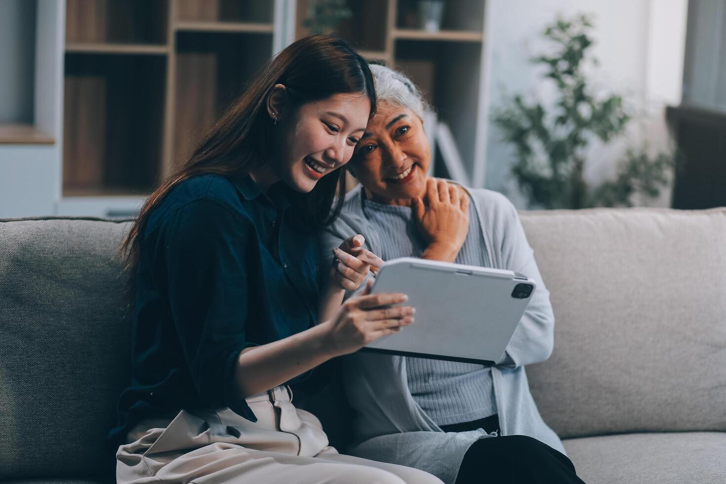 Happy grownup daughter showing content on tablet to mature mother. Two family generations women with digital computer gadget resting on couch together, watching videos, making call photo
