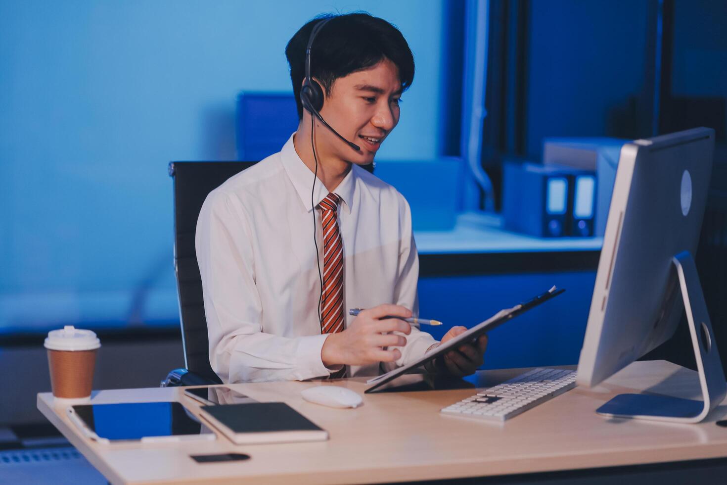 Happy smiling Asian man work at call center service desk consultant with teammates at night, customer service executive with microphone headset use computer for supporting in late night office. photo