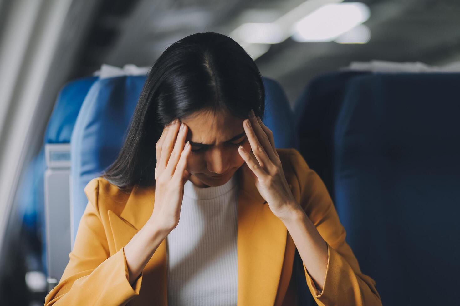 Young asian chinese airplane passenger woman experiencing discomfort, Airplane headache. Worried female passenger holding head, feeling unwell Anxiety and fear in the skies. Stressful travel moments. photo