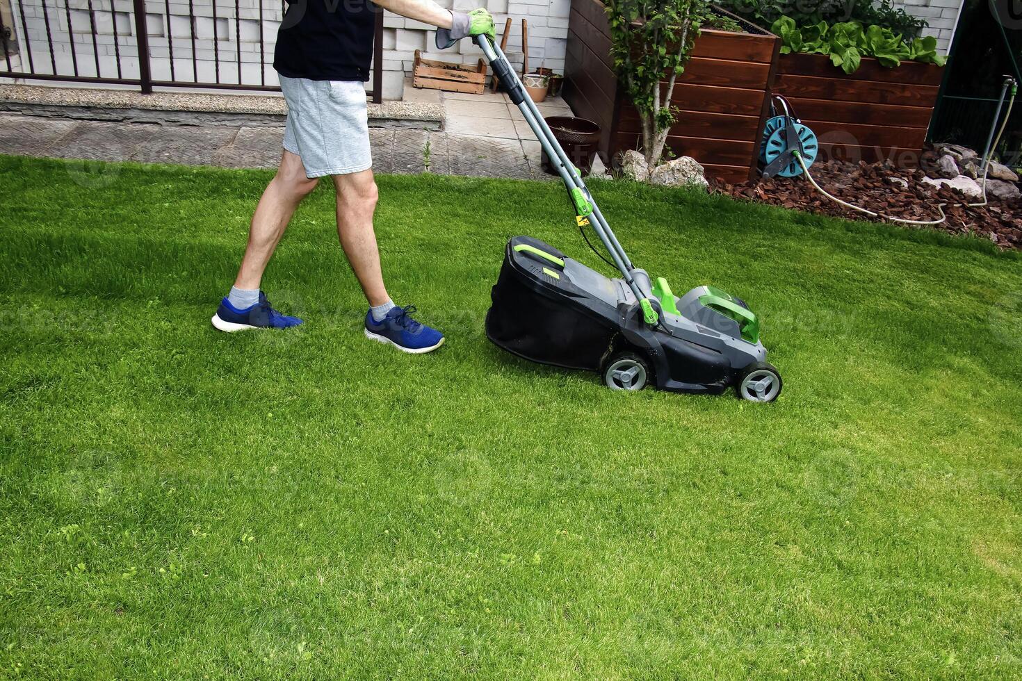A young man is mowing a lawn with a lawn mower in his beautiful green floral summer garden photo