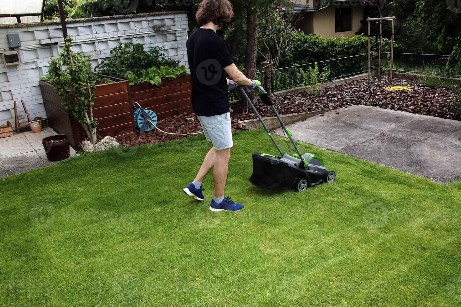 A young man is mowing a lawn with a lawn mower in his beautiful green floral summer garden photo