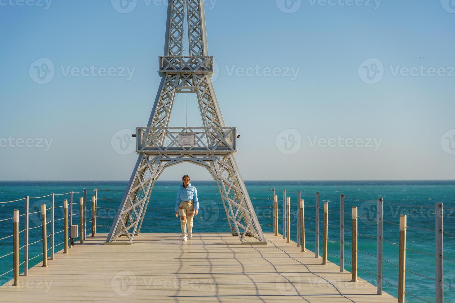 Large model of the Eiffel Tower on the beach. A woman walks along the pier towards the tower, wearing a blue jacket and white jeans. photo