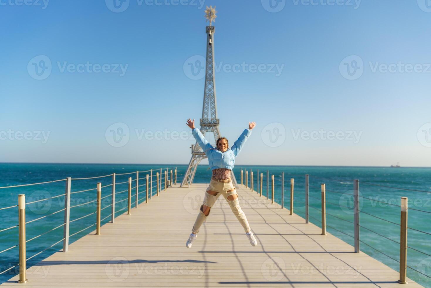 Large model of the Eiffel Tower on the beach. A woman walks along the pier towards the tower, wearing a blue jacket and white jeans. photo