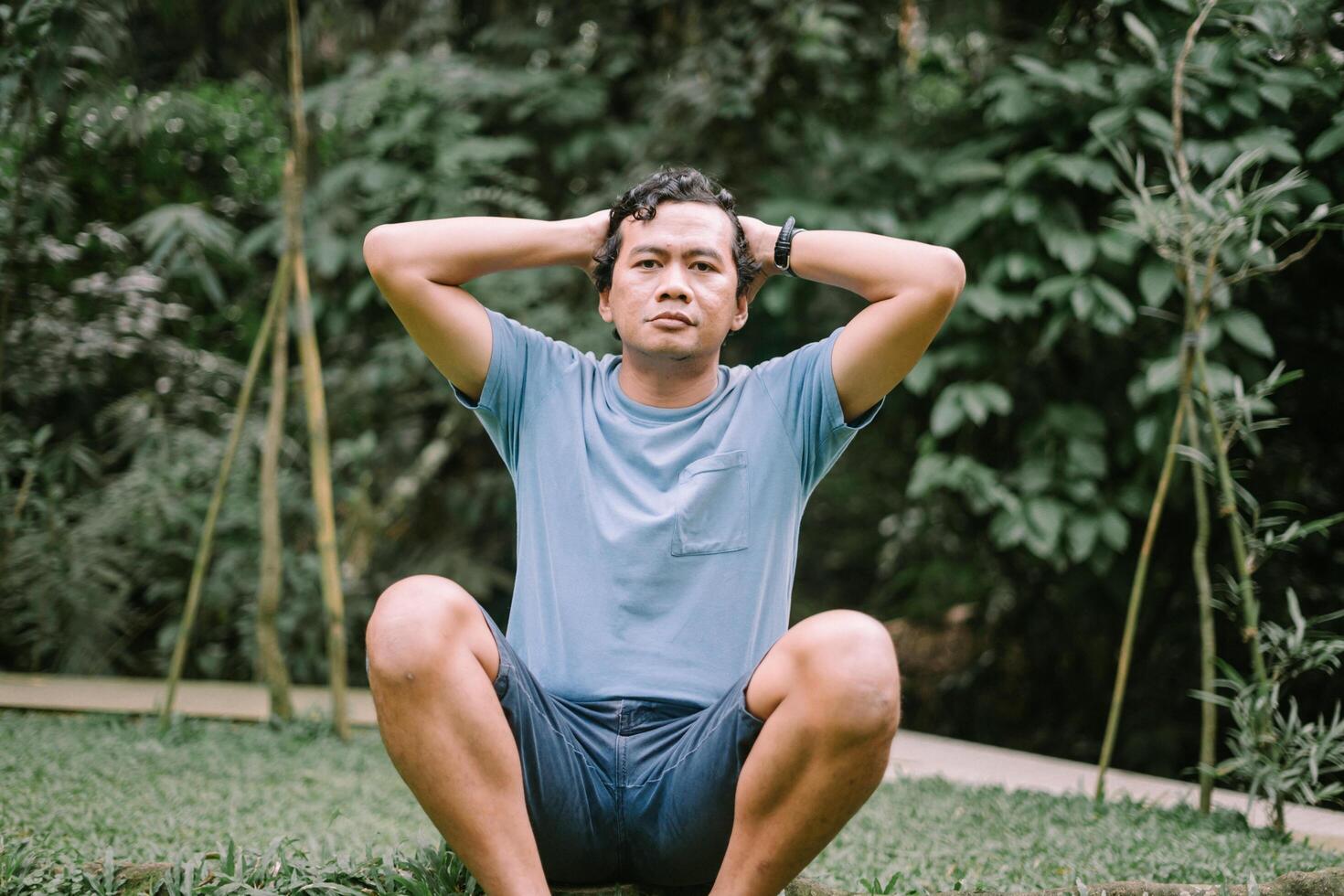 A Southeast Asian man sits on the ground with his hands on his head, expressing stress or deep thought, set against a natural backdrop photo