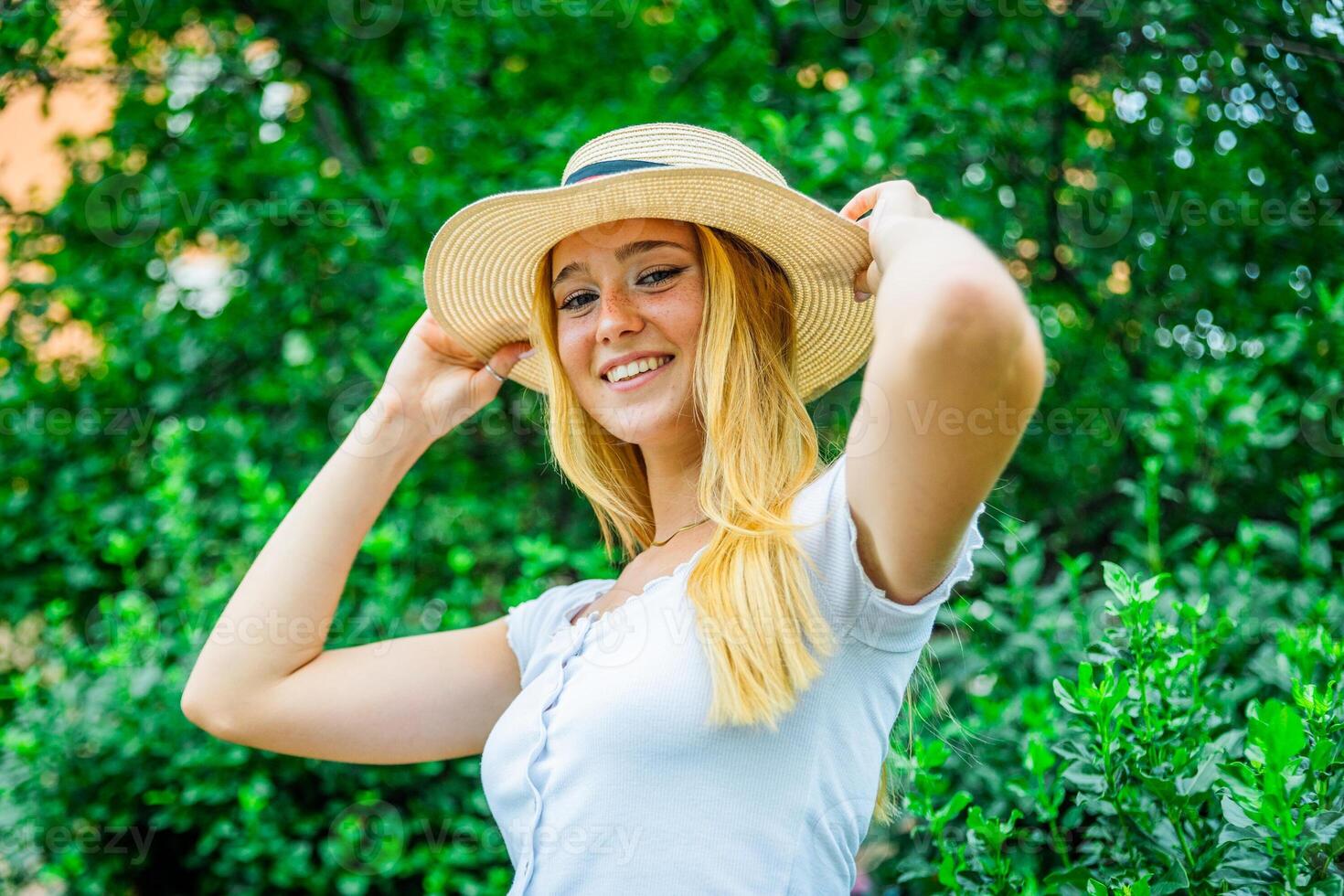 A smiling woman holds her summer hat while standing in nature photo