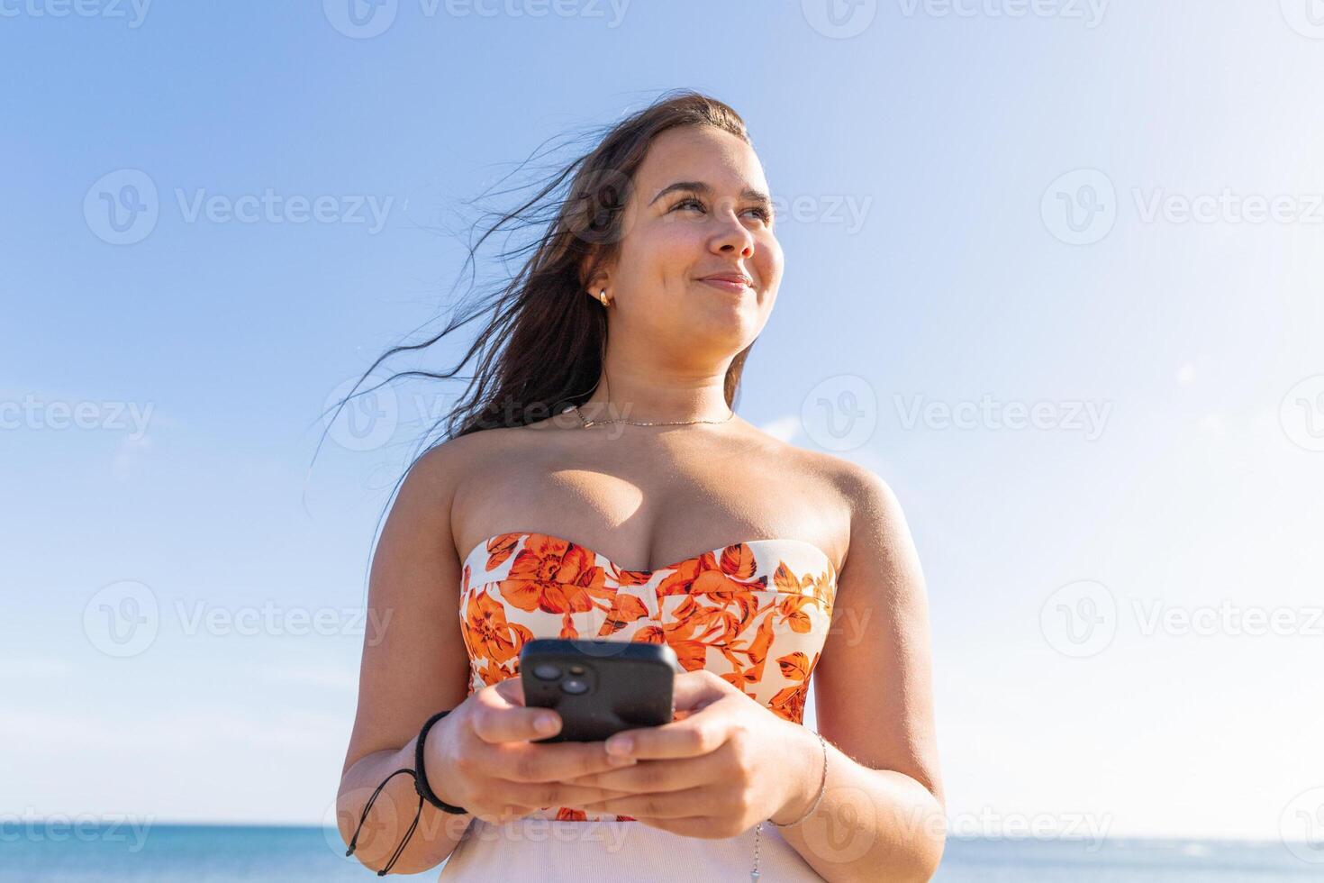 A teenage girl uses her smartphone happy while standing by the sea with the clear sky photo