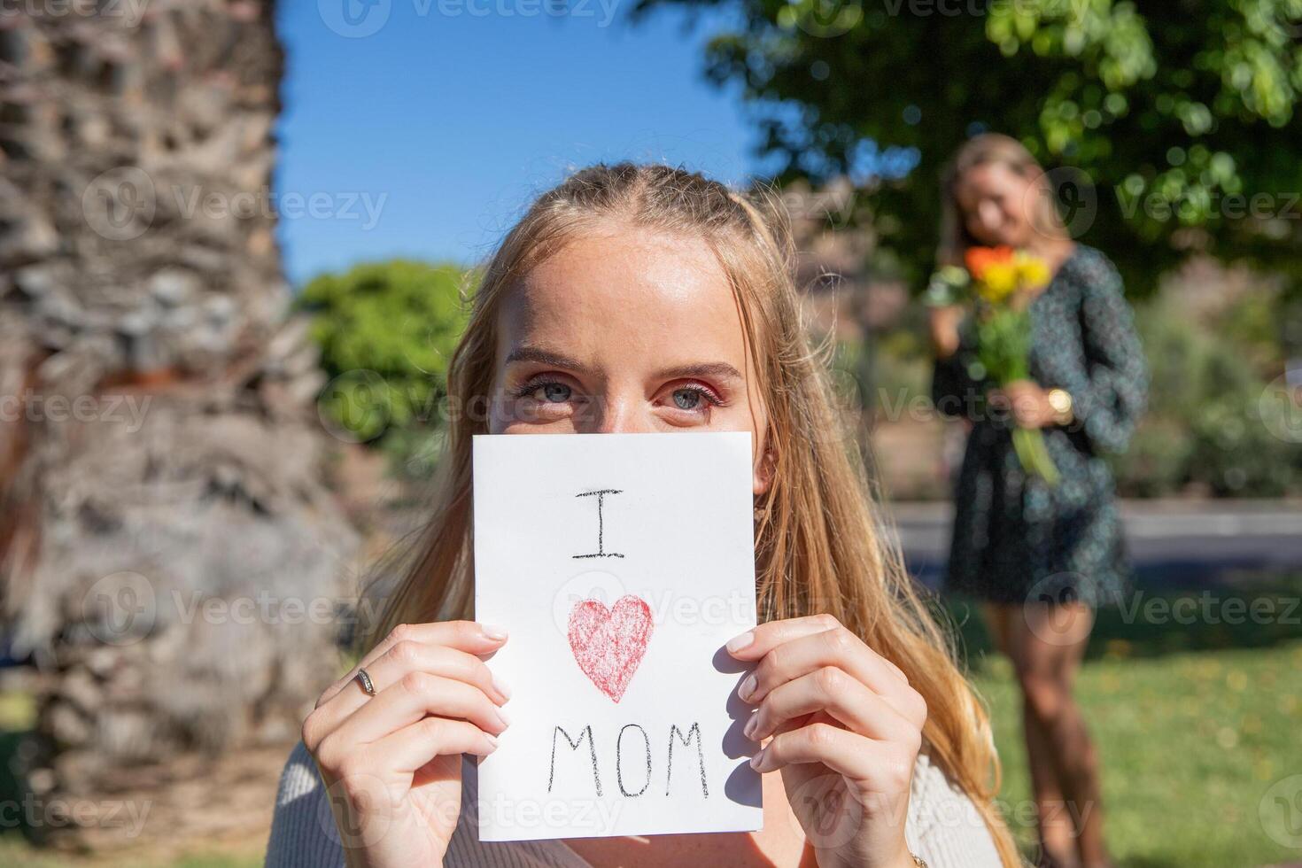 Daughter is holding card that says I Love mom with her mother behind with flowers received as gift photo