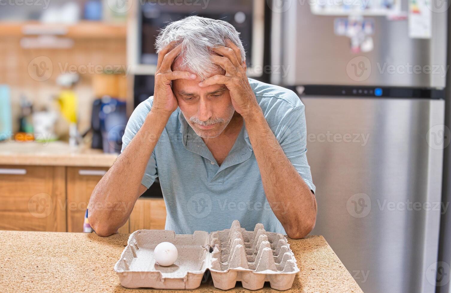 A desperate elderly gray-haired man grabbed his head, looking at the last chicken egg. The front is empty packaging. photo