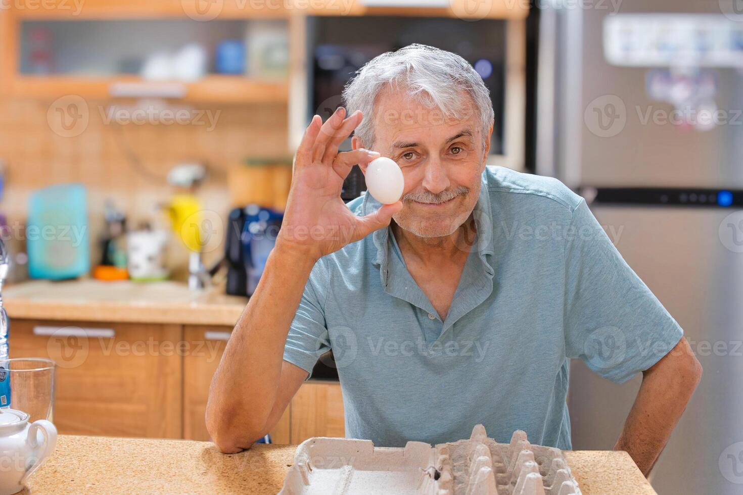 A smiling elderly gray-haired man, against the backdrop of the kitchen, holds the last chicken egg in his hand. The front is empty packaging. Looking straight into the camera. photo