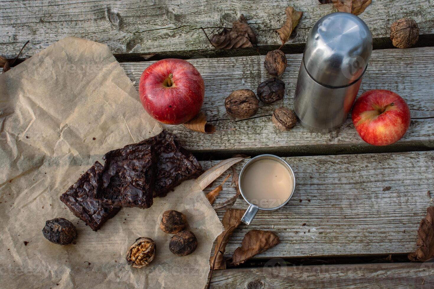 Piece of handmade brownie cake on the baking paper, metal thermos and mug with hot milk coffee, tee or cacao, red apple, nuts. Picnic on the wooden pier or terrace. Lunch break. Flat lay. Top view photo