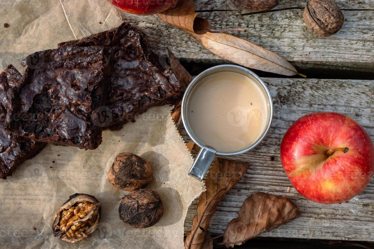 Close up of handmade brownie cake pieces on the baking paper, metal mug with hot milk coffee, tee or cacao, red apple, nuts. Picnic on the wooden pier or terrace. Lunch break. Flat lay. Top view photo