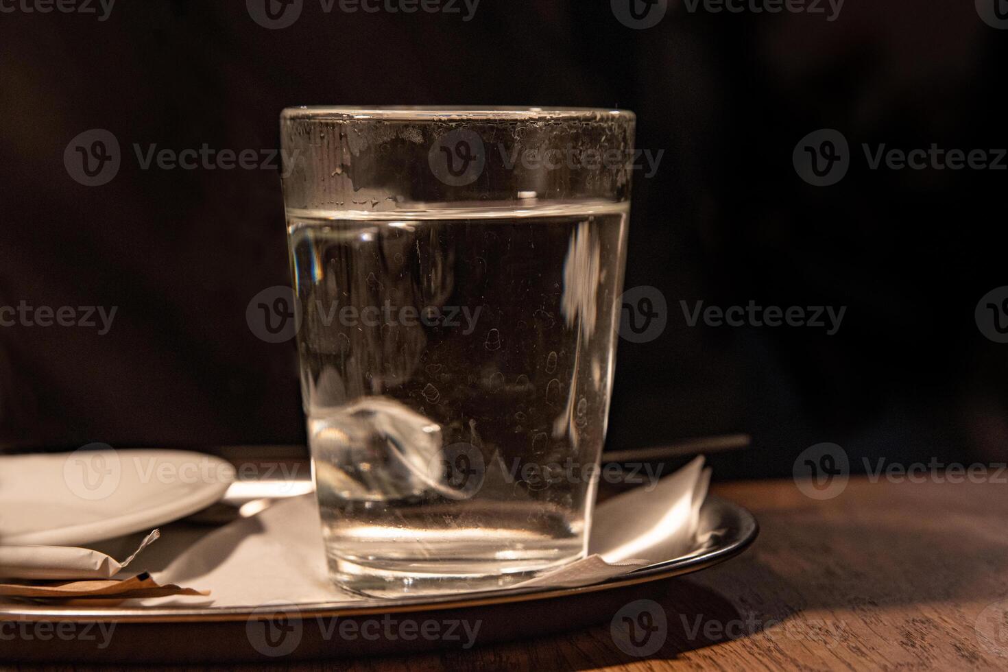 Close up of glass with hot water served on the white plate with napkin, sugar pack and tee bag on the wooden table in Cafe. Dark background. Drink concept. Tea cup. Selective focus. Copy space photo