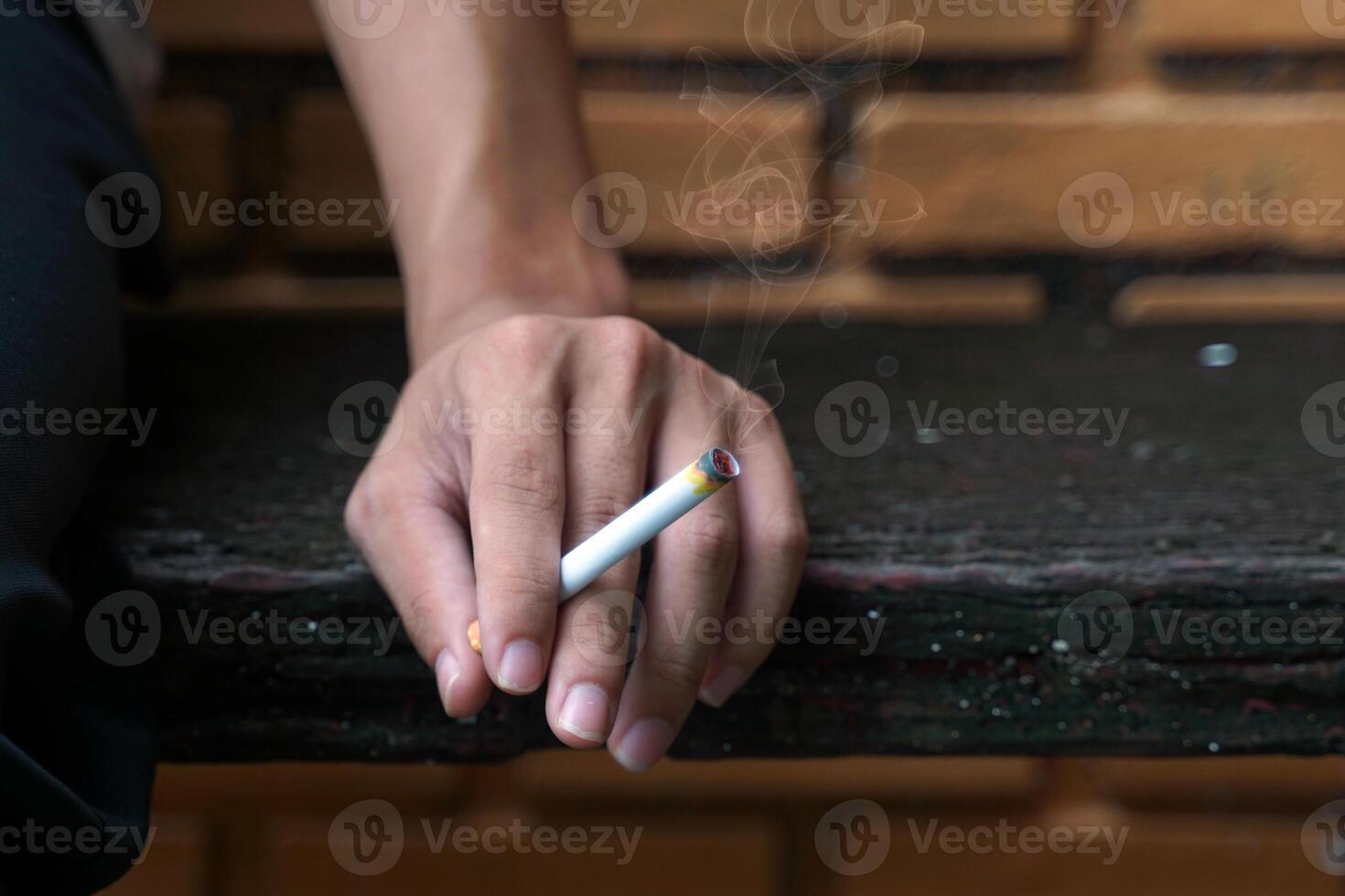 An Asian man's hand holds a lit cigarette and rests it on the wooden floor as he sits alone smoking in the smoking corner of his house. photo