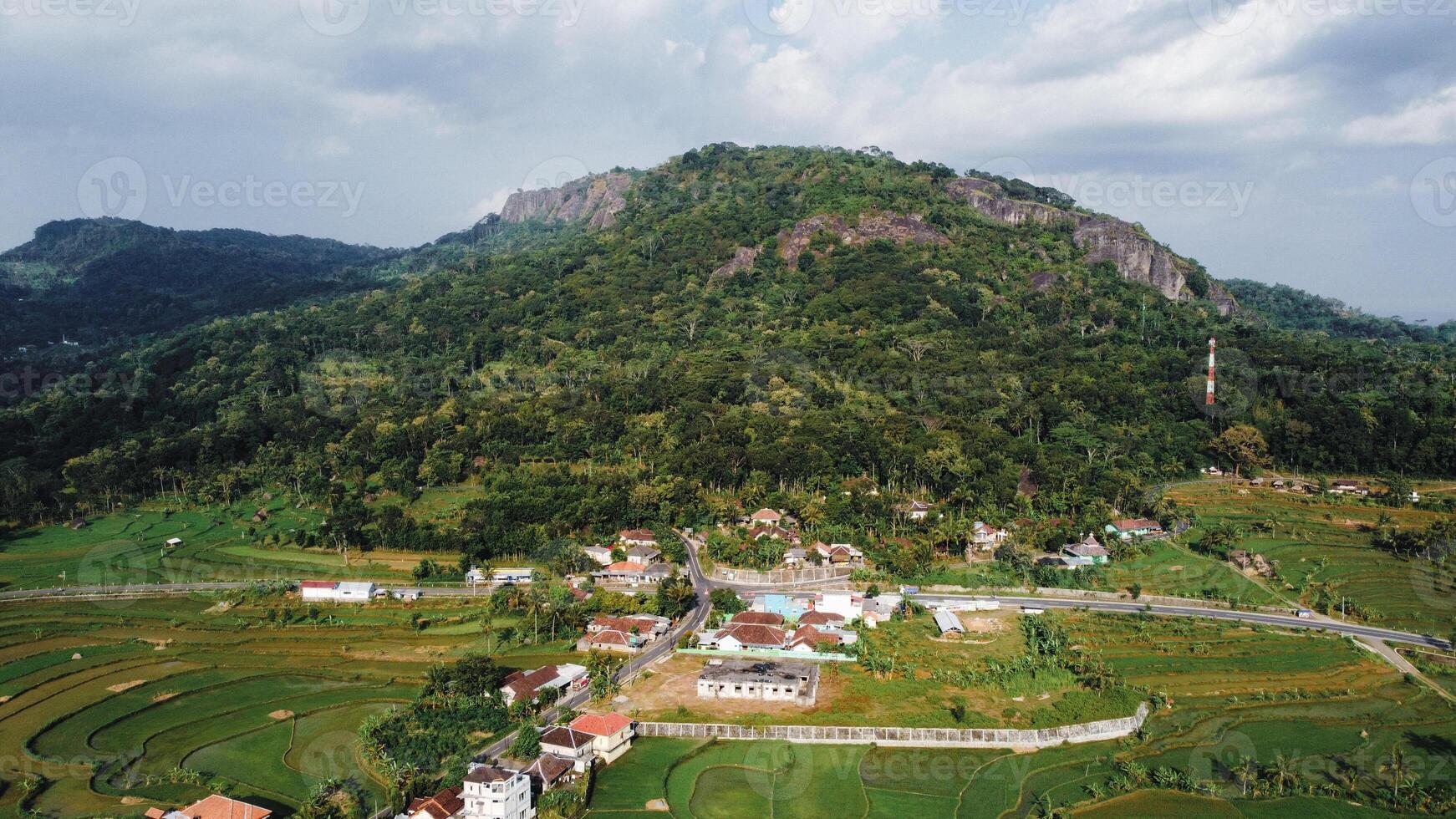 Panorama of the Nglanggeran Ancient Volcano ecotourism area in the afternoon using a drone. Aerial photo of an ancient stone hill covered with lush trees and surrounded by rice fields and countryside