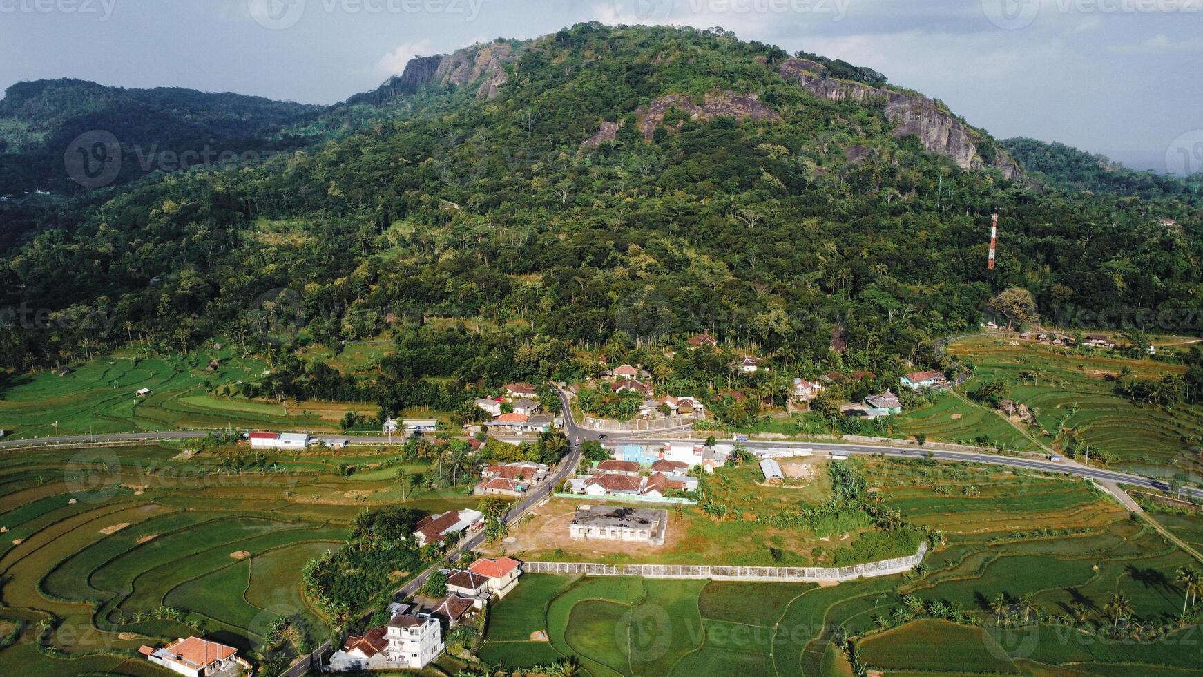 Panorama of the Nglanggeran Ancient Volcano ecotourism area in the afternoon using a drone. Aerial photo of an ancient stone hill covered with lush trees and surrounded by rice fields and countryside