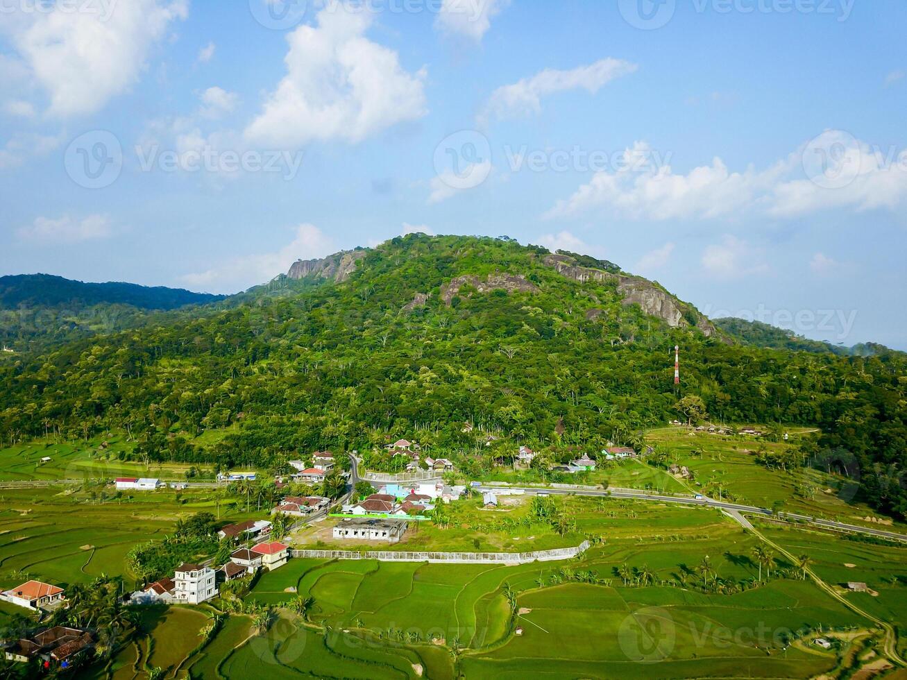 Panorama of the Nglanggeran Ancient Volcano ecotourism area in the afternoon using a drone. Aerial photo of an ancient stone hill covered with lush trees and surrounded by rice fields and countryside