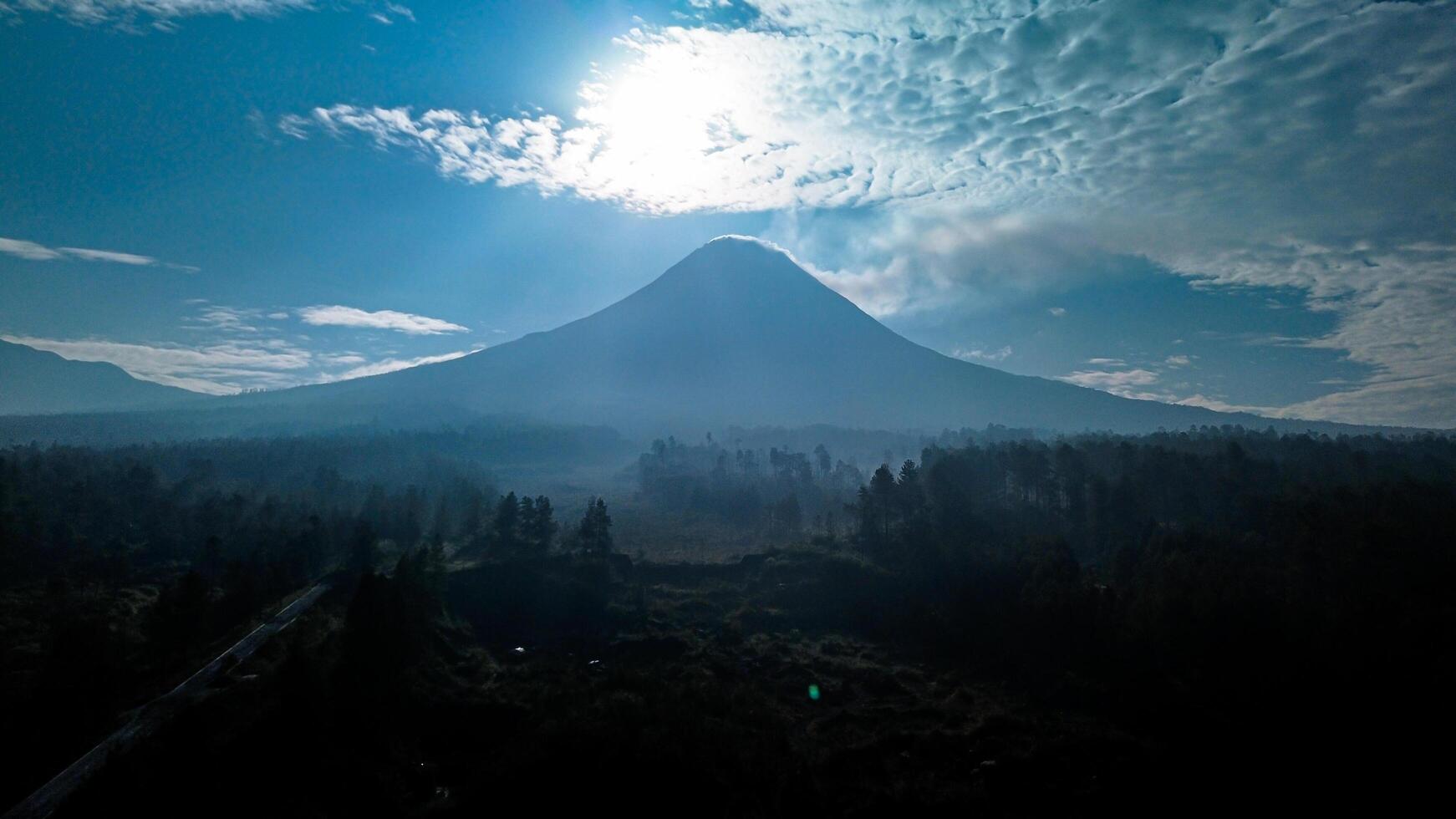 Landscape of Mount Merapi from a distance of 7 km before midday and the sun is directly behind the mountains. Aerial panorama of Mount Merapi during the day with white clouds around it photo