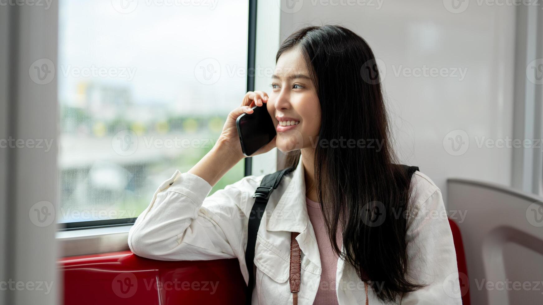 A happy Asian woman is looking at the view outside and talking on the phone while taking a sky train photo