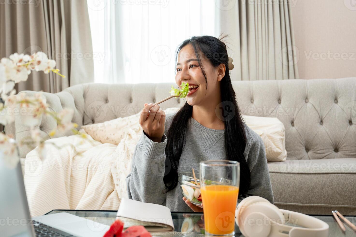 A charming Asian woman is enjoying eating healthy salad at a coffee table in the living room. photo